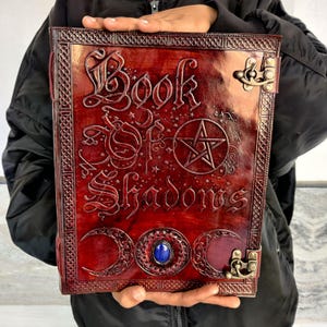 May include: A burgundy leather-bound book with the embossed title "Book of Shadows". The cover features a pentagram, crescent moons, and a central blue gemstone. Decorative metal clasps secure the book.