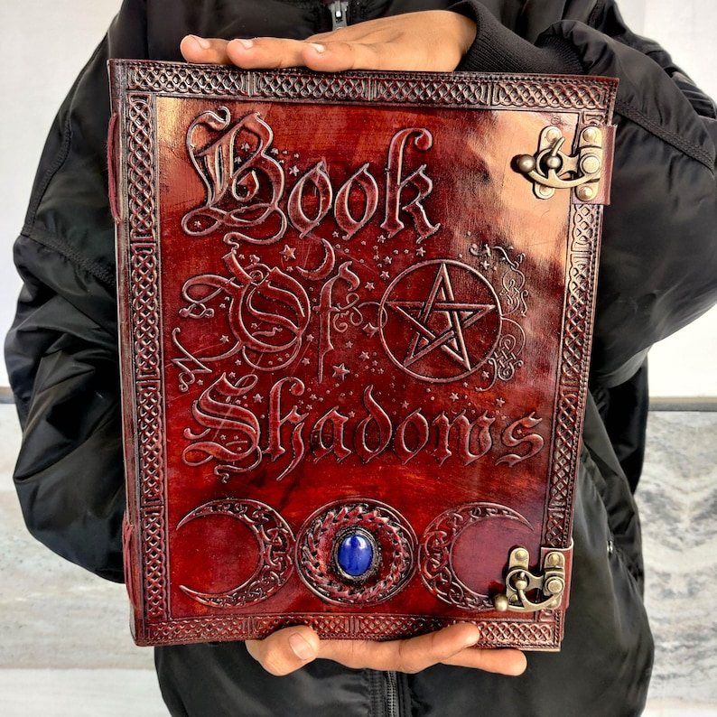 May include: A burgundy leather-bound book with the embossed title "Book of Shadows". The cover features a pentagram, crescent moons, and a central blue gemstone. Decorative metal clasps secure the book.