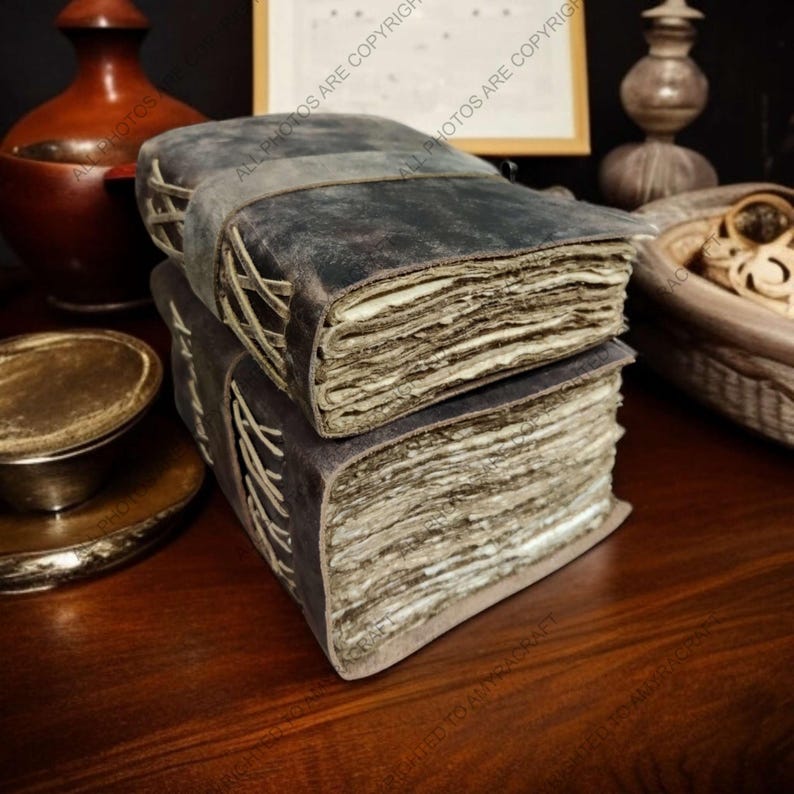 May include: Two stacked, antique-style journals with dark gray leather covers and aged, deckle-edged pages. The spines are hand-stitched with light-colored thread, and a leather strap secures the top journal. The journals rest on a dark wooden surface.