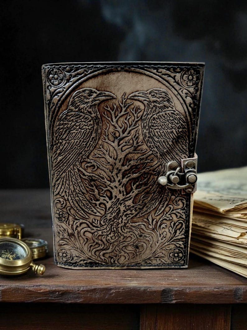 Puede incluir: Un diario de cuero marr&oacute;n con un dise&ntilde;o en relieve detallado que presenta dos cuervos enfrentados sobre un &aacute;rbol. El diario tiene un cierre de metal y patrones florales decorativos.