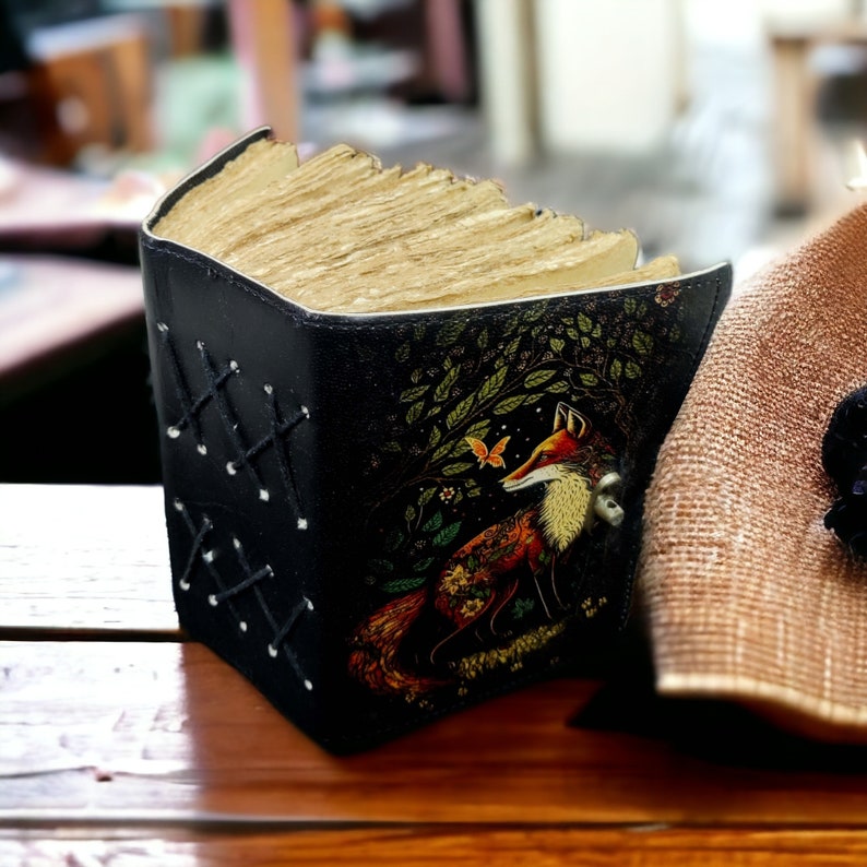 May include: A black book cover with a fox illustration, featuring a forest scene with a butterfly. The cover has a laced design on the side. The book's pages are aged and yellowed, giving it a vintage look.