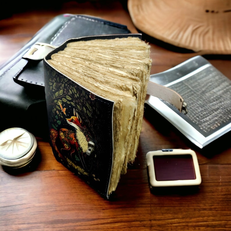 May include: A handcrafted journal with a leather cover depicting a fox. The journal has aged, textured pages and a leather bookmark. Other items on the wooden surface include a compass, a wallet, and a small notepad.