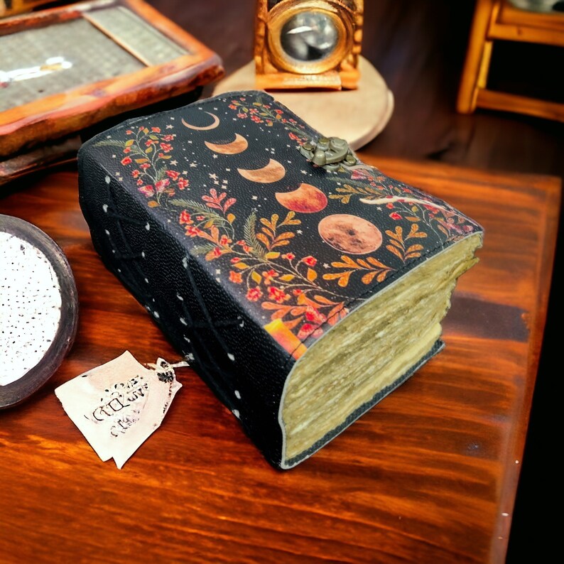 Puede incluir: Un diario encuadernado en cuero negro con un dise&ntilde;o floral y lunar. El diario tiene un aspecto desgastado y vintage y est&aacute; cerrado. El diario est&aacute; sobre una superficie de madera.