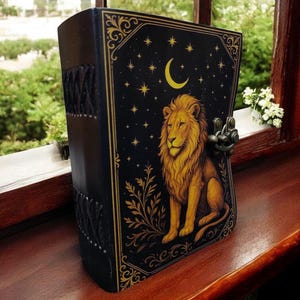 May include: A black leather-bound journal with a gold lion illustration on the cover. The cover also features a crescent moon, stars, and decorative gold filigree. The journal is secured with a metal clasp and sits on a wooden surface.