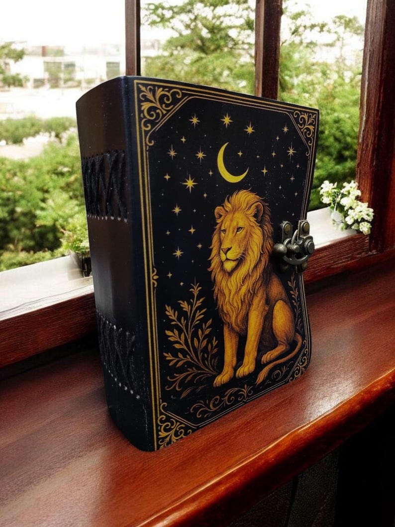May include: A black leather-bound journal with a gold lion illustration on the cover. The cover also features a crescent moon, stars, and decorative gold filigree. The journal is secured with a metal clasp and sits on a wooden surface.