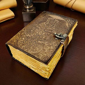 May include: A brown leather journal with an embossed design of two birds and foliage. The journal has a metal clasp and a leather strap closure. The pages have a yellowed, aged appearance. The journal is on a dark wooden surface.