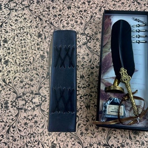 May include: A black leather-bound journal with cross-stitched detailing sits next to a boxed calligraphy set. The set includes a black feather quill, ink, and several nibs. The quill features a gold-colored ornate holder and a small bottle of ink.