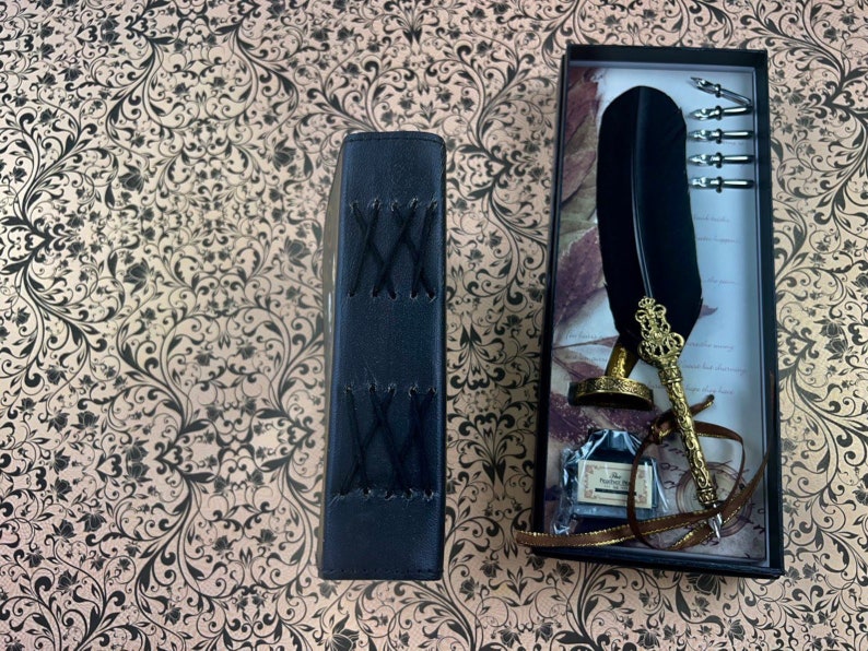 May include: A black leather-bound journal with cross-stitched detailing sits next to a boxed calligraphy set. The set includes a black feather quill, ink, and several nibs. The quill features a gold-colored ornate holder and a small bottle of ink.