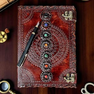 May include: A handcrafted, reddish-brown leather journal with intricate embossed designs and a decorative gemstone accent. A black fountain pen rests on the journal. The journal has a metal clasp closure. Other antique items are in the background.