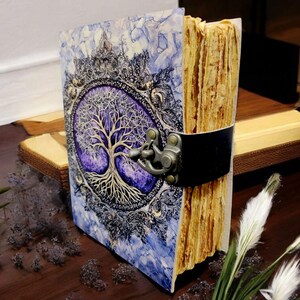 Op de afbeelding: Een handgemaakt dagboek met een levensboommotief. De omslag toont een gedetailleerde boomillustratie met ingewikkelde takken en wortels, tegen een paarse en blauwe achtergrond. Het dagboek is vastgezet met een zwarte leren riem en metalen sluiting.