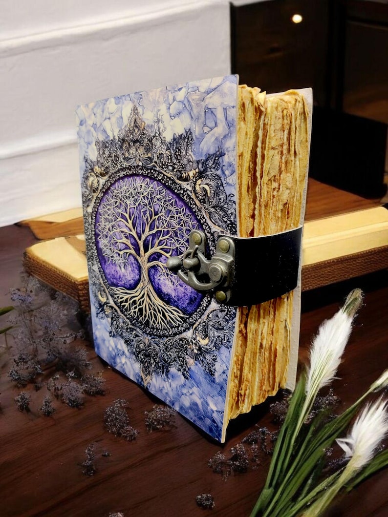 Op de afbeelding: Een handgemaakt dagboek met een levensboommotief. De omslag toont een gedetailleerde boomillustratie met ingewikkelde takken en wortels, tegen een paarse en blauwe achtergrond. Het dagboek is vastgezet met een zwarte leren riem en metalen sluiting.
