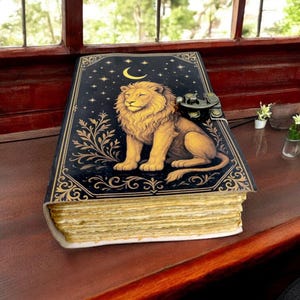 May include: A decorative journal with a lion illustration. The cover is black with gold accents, featuring a lion, crescent moon, and stars. The journal has a metal clasp and aged-looking pages. It rests on a dark wooden surface near a window.