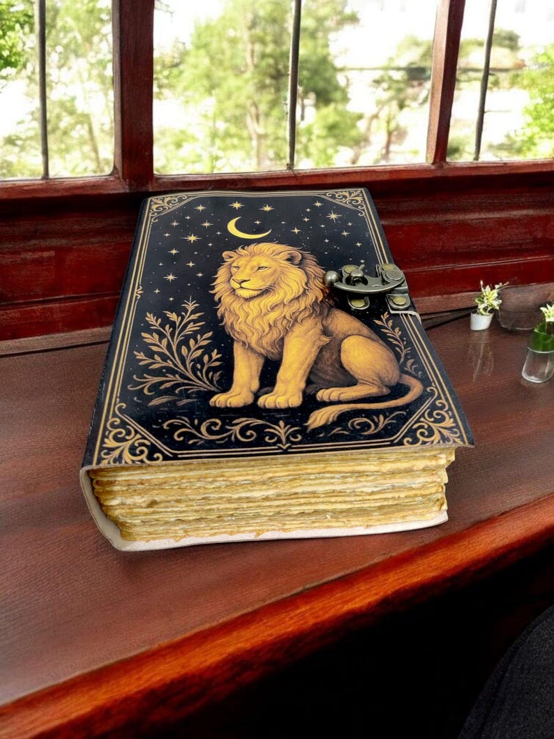 May include: A decorative journal with a lion illustration. The cover is black with gold accents, featuring a lion, crescent moon, and stars. The journal has a metal clasp and aged-looking pages. It rests on a dark wooden surface near a window.