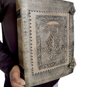May include: A large, beige leather-bound journal with intricate embossed designs. The cover features a central panel with two birds flanking a tree, surrounded by floral and geometric patterns. Metal clasps secure the journal.