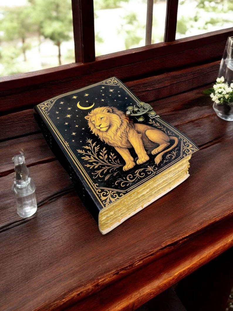 May include: A decorative journal with a lion design. The cover is black with gold accents, a crescent moon, and stars. The lion is golden, and the pages have a gold edge. The journal has a metal clasp and is on a wooden surface.