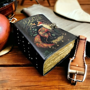 May include: A black leather journal featuring a fox illustration in a forest setting. The journal has a metal clasp and is studded along the spine. The pages have a vintage appearance, and the cover art displays vibrant colours.