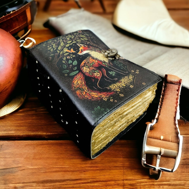 May include: A black leather journal featuring a fox illustration in a forest setting. The journal has a metal clasp and is studded along the spine. The pages have a vintage appearance, and the cover art displays vibrant colours.