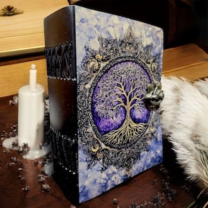 Op de afbeelding: Een decoratief boek met een levensboomontwerp. De kaft heeft een gedetailleerde illustratie van een gouden boom tegen een paarse achtergrond, omlijst door sierlijke zwarte en gouden accenten. De rug is zwart met decoratieve stiksels. Het boek staat op een houten oppervlak.