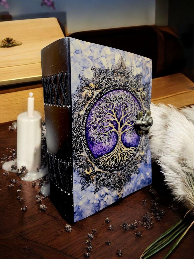 Op de afbeelding: Een decoratief boek met een levensboomontwerp. De kaft heeft een gedetailleerde illustratie van een gouden boom tegen een paarse achtergrond, omlijst door sierlijke zwarte en gouden accenten. De rug is zwart met decoratieve stiksels. Het boek staat op een houten oppervlak.