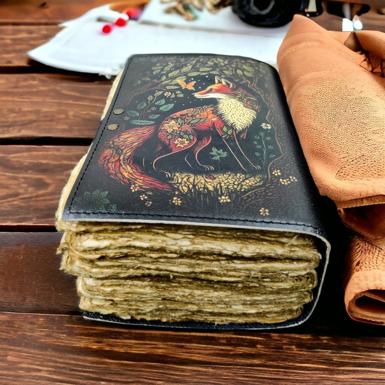 May include: A handcrafted journal with a black cover. The cover displays a detailed illustration of a fox in a forest, with orange, red, and cream tones, and green foliage. The journal has thick, aged pages.
