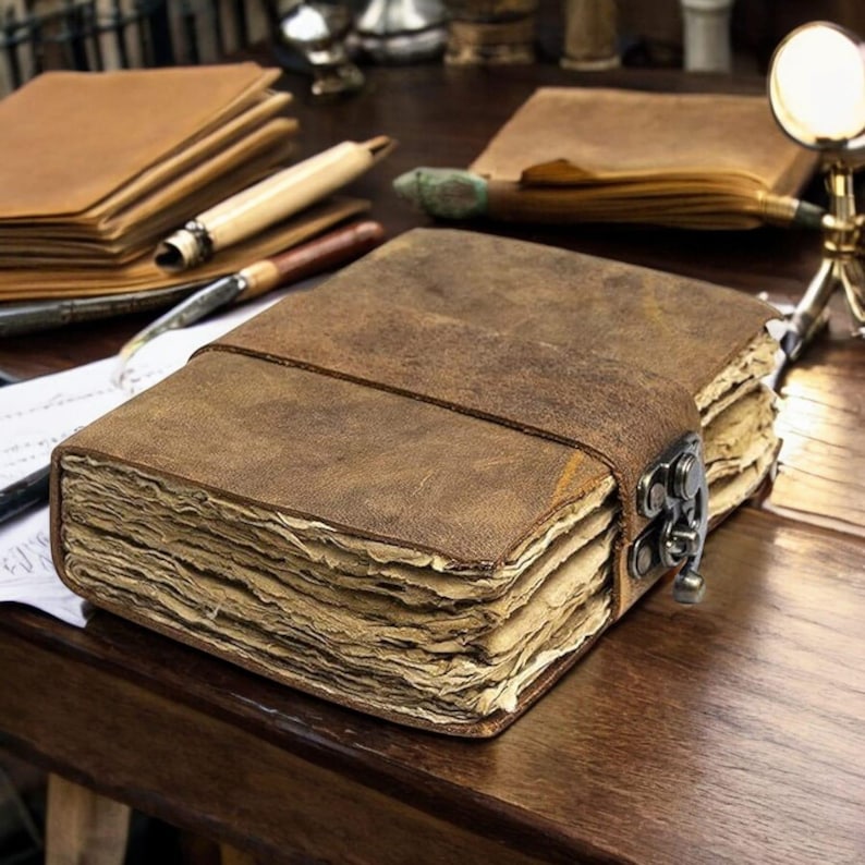 May include: A brown leather journal with a metal clasp and a strap closure. The journal has thick, aged pages and is sitting on a wooden surface. Other journals and writing instruments are in the background.