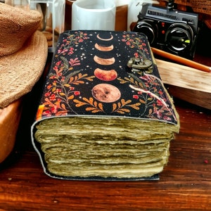 Puede incluir: Un diario encuadernado en cuero negro con un dise&ntilde;o floral y fases lunares. El diario tiene un cierre de lat&oacute;n y est&aacute; lleno de p&aacute;ginas en blanco.