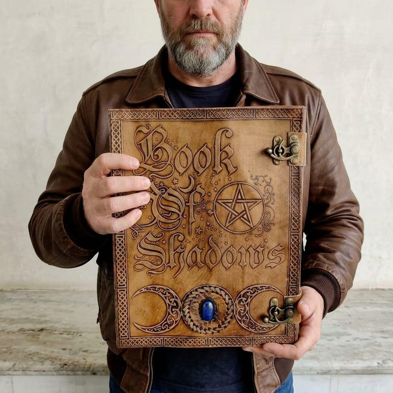 May include: A light brown leather-bound book with the title "Book of Shadows" embossed on the cover. The cover features a pentagram, crescent moons, and a blue gemstone. The book has metal clasps.