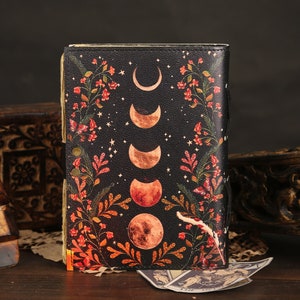 Puede incluir: Un diario de cuero negro con un dise&ntilde;o de fases lunares y flores. El diario tiene un aspecto desgastado y est&aacute; decorado con detalles dorados.