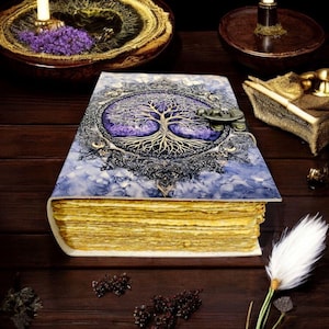 Op de afbeelding: Een handgemaakt notitieboek met een levensboomontwerp op de kaft. De kaft is een mix van blauw en paars met een sierlijke metalen sluiting. De pagina's hebben een gouden tint en het notitieboek ligt op een donker houten oppervlak.