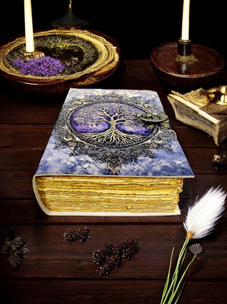 Op de afbeelding: Een handgemaakt notitieboek met een levensboomontwerp op de kaft. De kaft is een mix van blauw en paars met een sierlijke metalen sluiting. De pagina's hebben een gouden tint en het notitieboek ligt op een donker houten oppervlak.