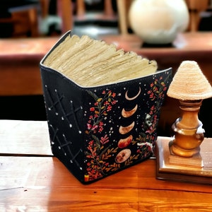 Puede incluir: Un diario de cuero negro con un dise&ntilde;o floral y lunar. El diario est&aacute; cerrado y tiene una pila de p&aacute;ginas en blanco en su interior.