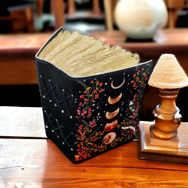 Puede incluir: Un diario de cuero negro con un dise&ntilde;o floral y lunar. El diario est&aacute; cerrado y tiene una pila de p&aacute;ginas en blanco en su interior.