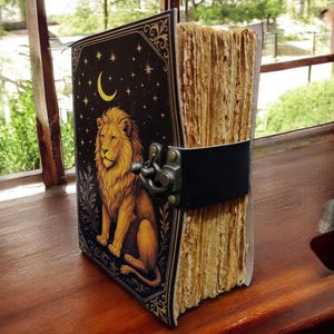 May include: A vintage-style journal with a lion illustration on the cover. The cover is black with gold accents, a crescent moon, and stars. The journal has a black leather strap and clasp closure. The pages are aged and textured.