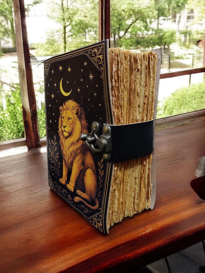 May include: A vintage-style journal with a lion illustration on the cover. The cover is black with gold accents, a crescent moon, and stars. The journal has a black leather strap and clasp closure. The pages are aged and textured.