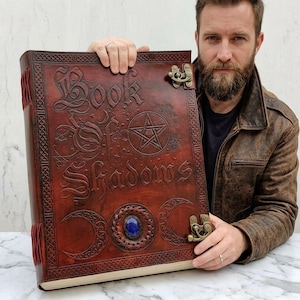 Op de afbeelding: Een groot, ledergebonden boek met de titel "Book of Shadows" in reliëf op de kaft. De kaft is een rijke, roodbruine kleur, met een pentagram, halve manen en ingewikkelde Keltische knoopwerkontwerpen. Het boek heeft een messing sluiting en een blauwe edelsteen.