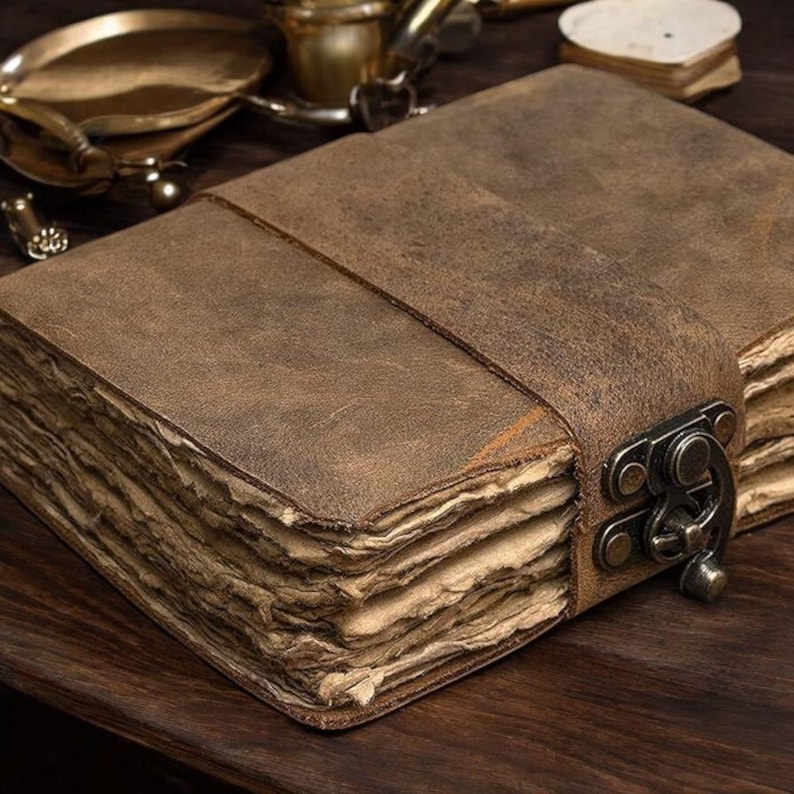 May include: A brown leather journal with a strap closure and metal clasp. The journal has aged, textured pages and a leather cover. The journal is closed with a leather strap and metal clasp. The journal is on a wooden surface.