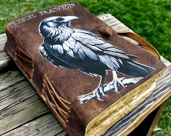 Könnte beinhalten: Ein braunes Lederjournal mit einer schwarzen Rabenillustration und dem Text "True Raven" auf dem Cover. Das Journal hat einen Lederriemenverschluss und eine ausgefranste Kante.