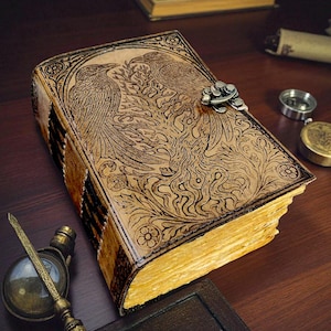 May include: A leather-bound journal with an embossed design of two birds and floral patterns. The journal has a metal clasp and aged, yellowed pages. The cover is a light brown color, and the edges of the pages are gilded.