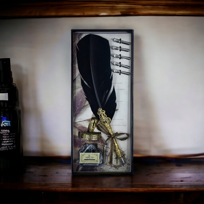 Op de afbeelding: Een vintage kalligrafieset met een grote zwarte veerkiel met een goudkleurige metalen penpunt en decoratieve accenten. De set bevat een kleine inktfles en verschillende penpunten, allemaal gepresenteerd in een rechthoekige doos.