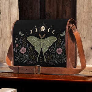 May include: A black messenger bag with a brown leather strap and trim. The front flap features a large, pale green moth centred among botanical illustrations of pink flowers, dark leaves, and a series of moon phases with small stars.