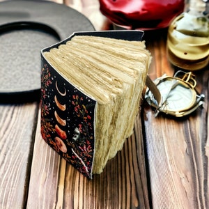 Puede incluir: Un diario encuadernado en cuero negro con un dise&ntilde;o floral y lunar. El diario est&aacute; abierto, revelando p&aacute;ginas en blanco.