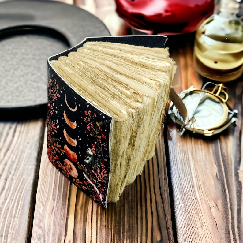 Puede incluir: Un diario encuadernado en cuero negro con un dise&ntilde;o floral y lunar. El diario est&aacute; abierto, revelando p&aacute;ginas en blanco.