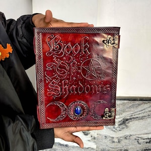 May include: A red leather-bound book titled "Book of Shadows" with embossed lettering and symbols. The cover includes a pentagram, crescent moons, and a blue gemstone. The book is held open, displaying the detailed design and craftsmanship.