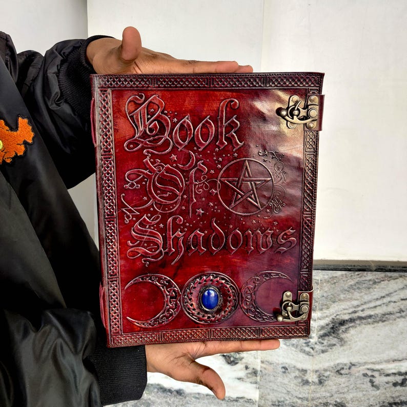 May include: A red leather-bound book titled "Book of Shadows" with embossed lettering and symbols. The cover includes a pentagram, crescent moons, and a blue gemstone. The book is held open, displaying the detailed design and craftsmanship.
