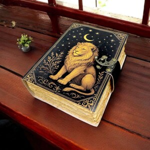 May include: A decorative journal with a black cover featuring a golden lion illustration, a crescent moon, and stars. The journal has a clasp closure and aged-looking pages. The journal is resting on a wooden surface.