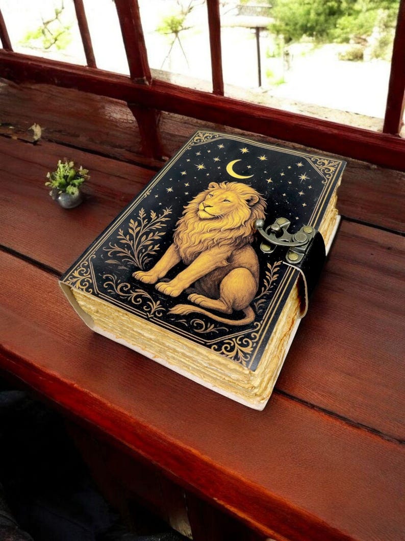 May include: A decorative journal with a black cover featuring a golden lion illustration, a crescent moon, and stars. The journal has a clasp closure and aged-looking pages. The journal is resting on a wooden surface.