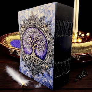 Op de afbeelding: Een decoratief notitieboek met een levensboomontwerp. De kaft heeft een gouden boom op een paarse achtergrond, omlijst door een sierlijke gouden en zwarte rand. De rug is zwart met gestikte details. Het notitieboek ligt op een houten oppervlak.