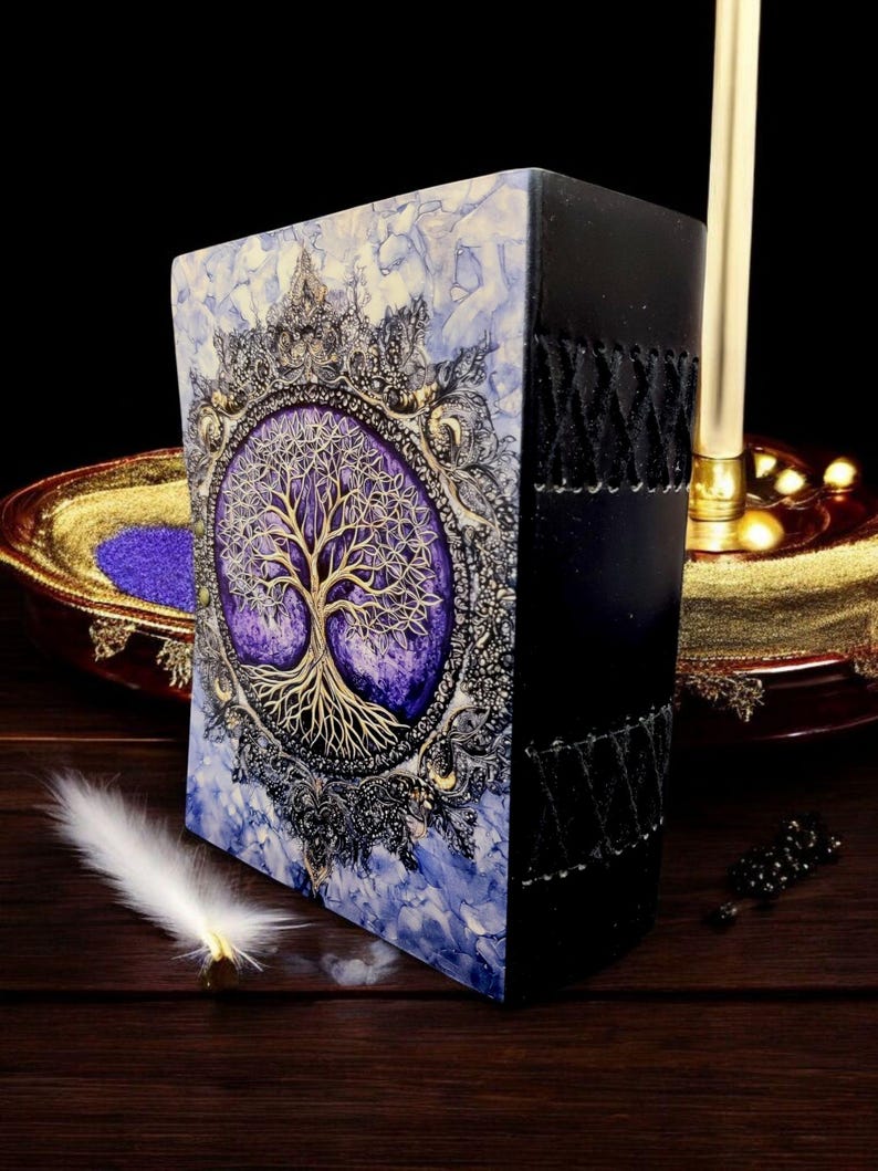 Op de afbeelding: Een decoratief notitieboek met een levensboomontwerp. De kaft heeft een gouden boom op een paarse achtergrond, omlijst door een sierlijke gouden en zwarte rand. De rug is zwart met gestikte details. Het notitieboek ligt op een houten oppervlak.