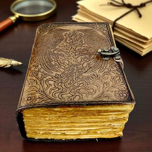 May include: A brown leather journal with an embossed design featuring two birds and floral patterns. The journal has a metal clasp and aged, yellowed pages. A magnifying glass and stationery are in the background.