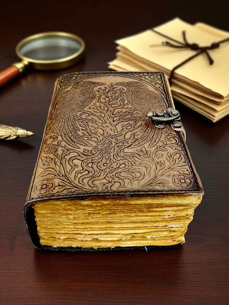 May include: A brown leather journal with an embossed design featuring two birds and floral patterns. The journal has a metal clasp and aged, yellowed pages. A magnifying glass and stationery are in the background.